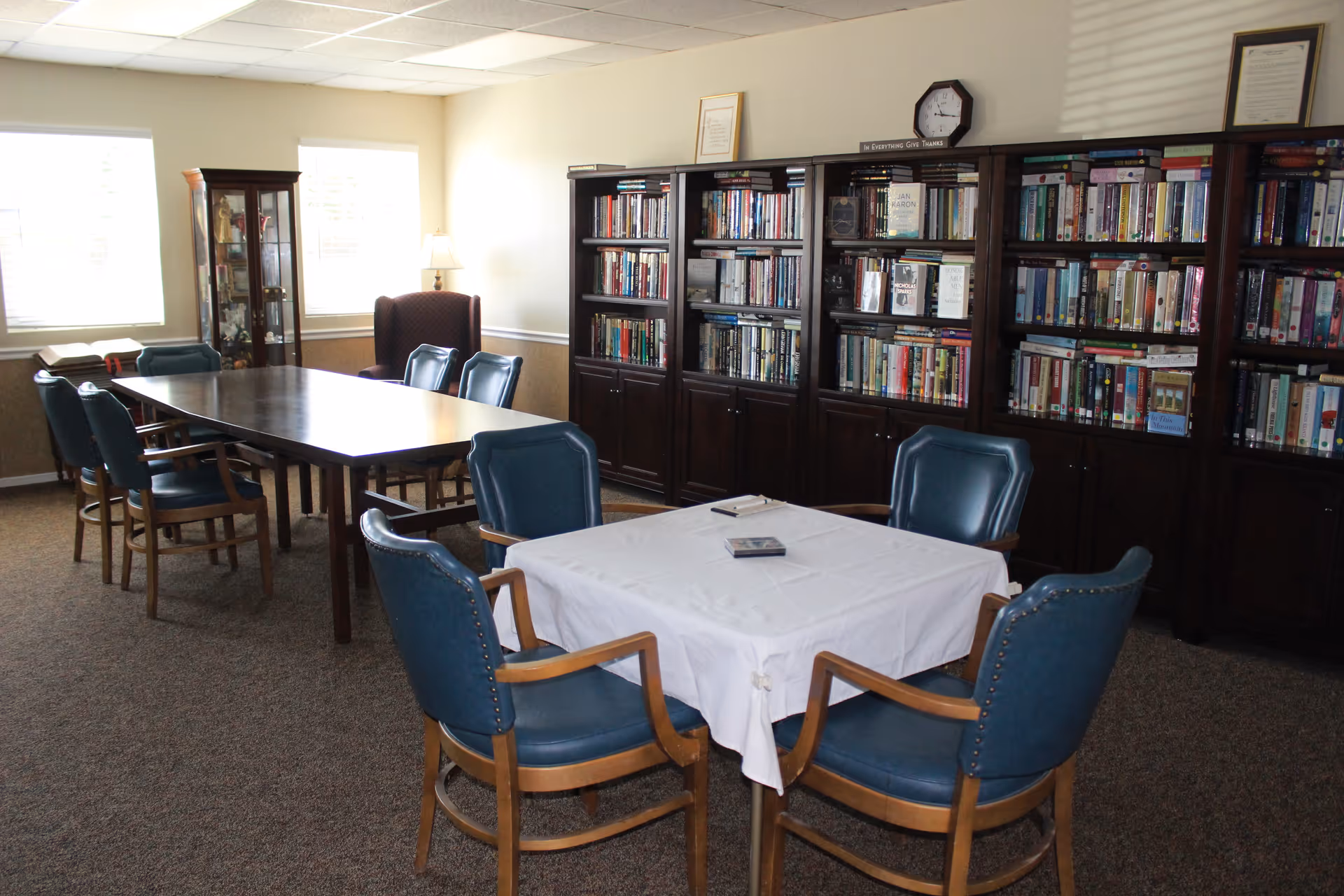 A quiet room with two tables surrounded by blue cushioned chairs. One table is covered with a white tablecloth and has a few items on it. The other table is wooden and longer, with several chairs around it. Along the back wall are large dark wooden bookshelves filled with books. There is a glass display cabinet and a lamp near two windows letting in natural light.
