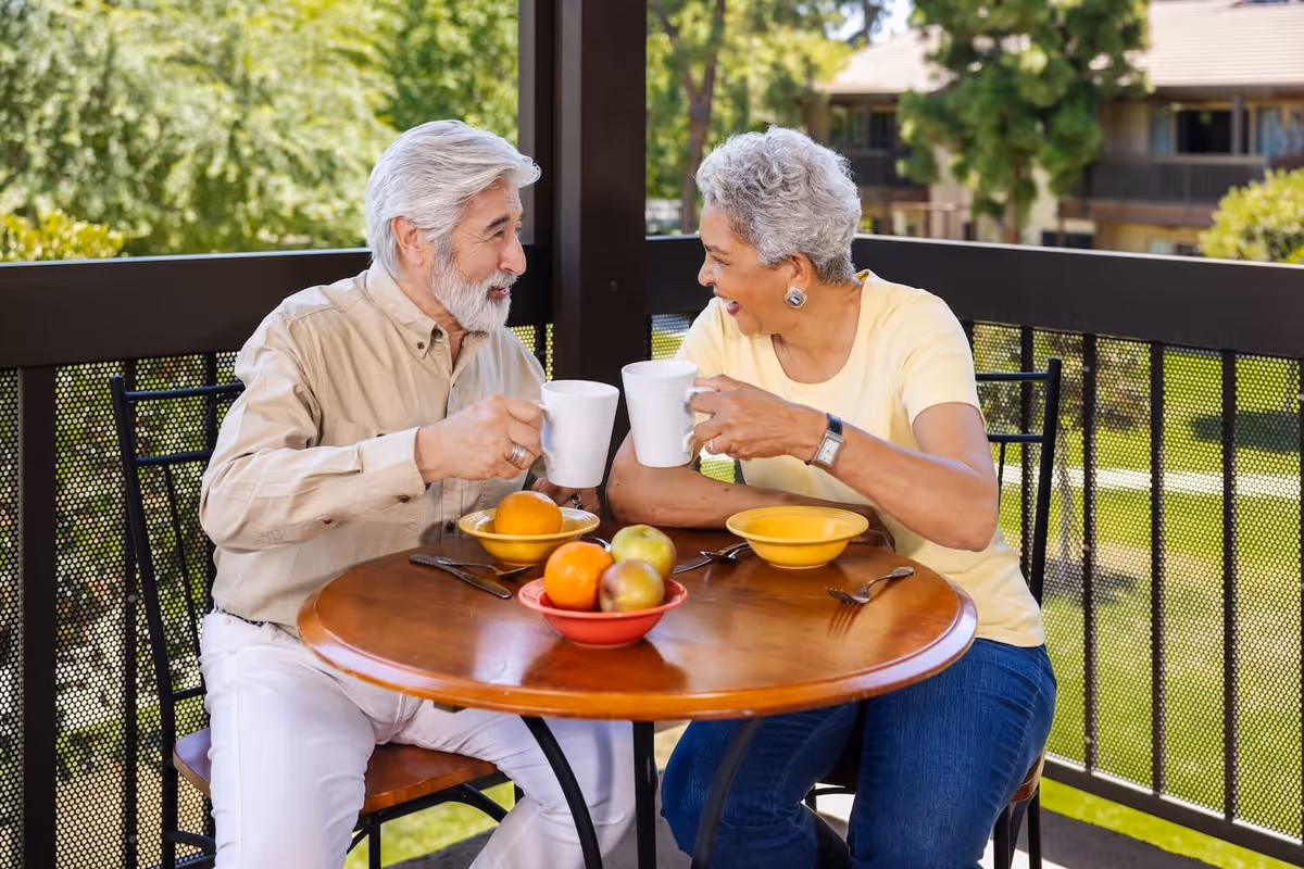 An elderly man and woman sitting at a round wooden table on a balcony, clinking white mugs and smiling at each other. The table has bowls with fruit and utensils. Green trees and a building are visible in the background.