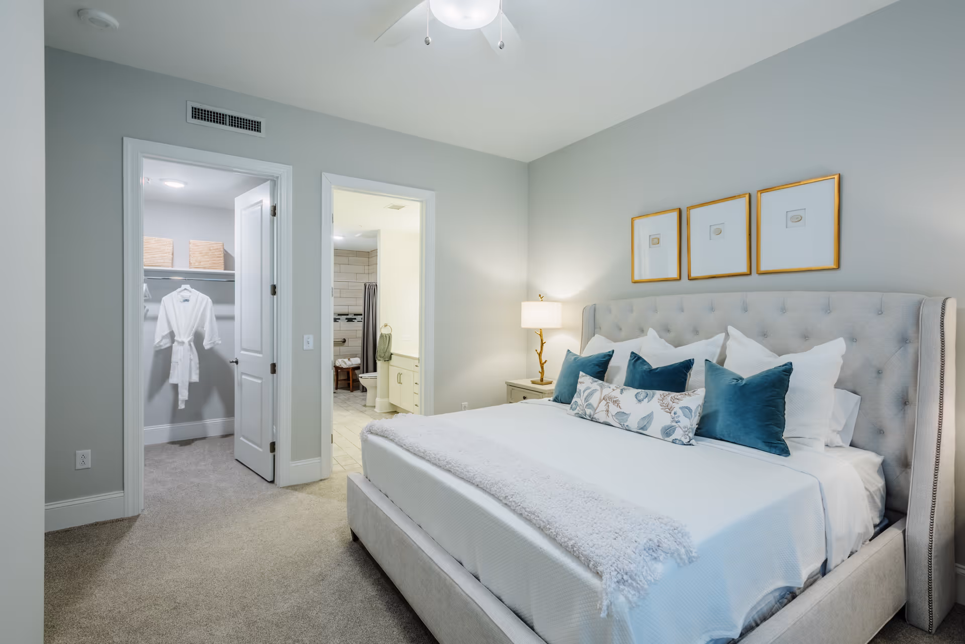 Well-lit bedroom with a tufted bed dressed in white and teal pillows, a nightstand lamp, and open doors to a walk-in closet and bathroom.