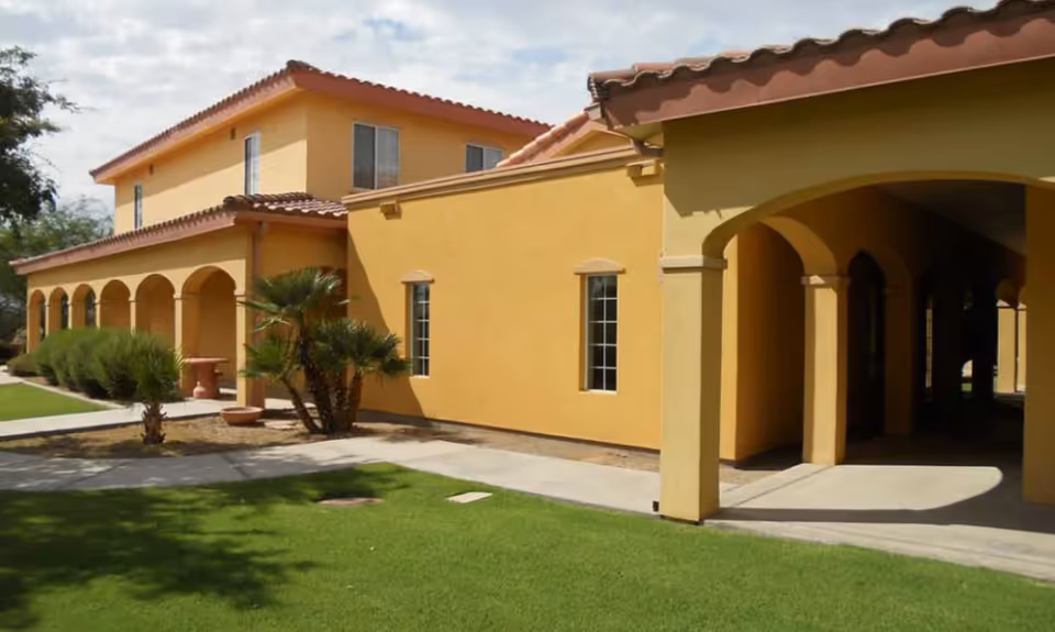 Exterior view of a yellow stucco building with a red tile roof, featuring arched walkways and windows, surrounded by green grass and small palm trees under a partly cloudy sky.