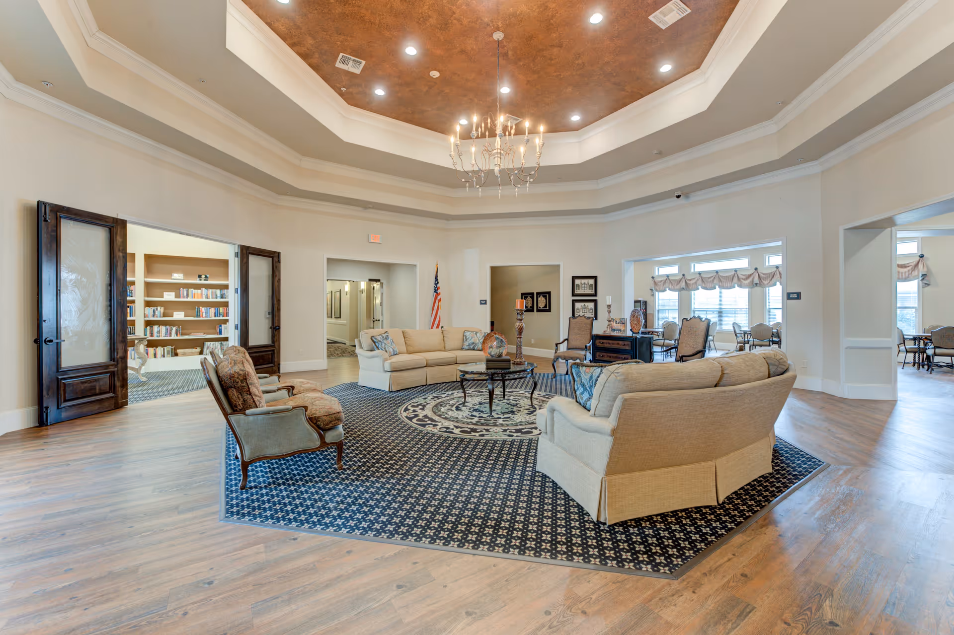 A spacious senior living facility common area with a high tray ceiling featuring a chandelier. The room has a patterned carpet area with beige sofas and armchairs arranged around a central coffee table. There are multiple doorways leading to other rooms, including a library with bookshelves and a dining area with tables and chairs. The floor is wood, and the walls are painted light beige.