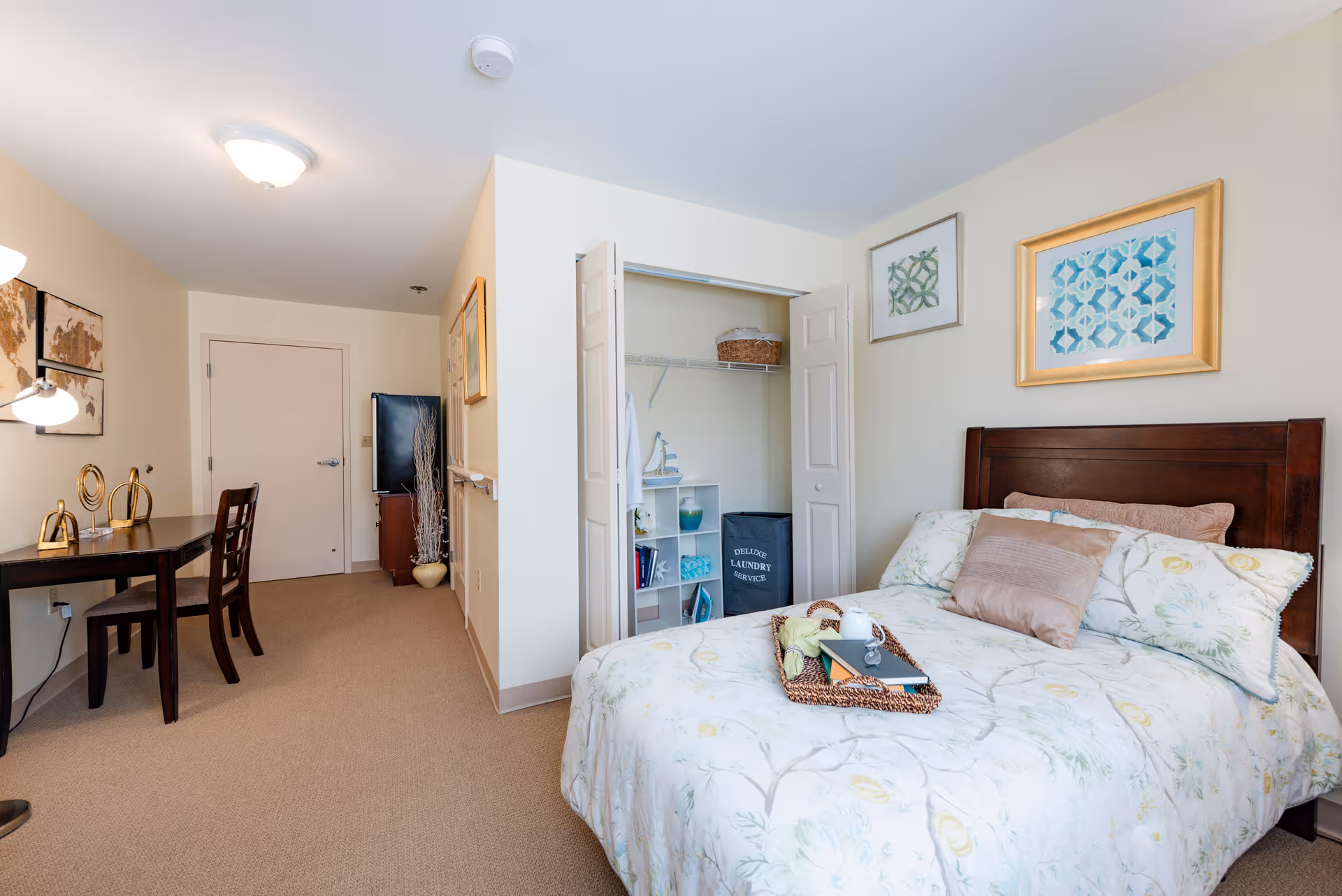 A furnished senior living bedroom with a bed, desk, open closet, and framed wall art.