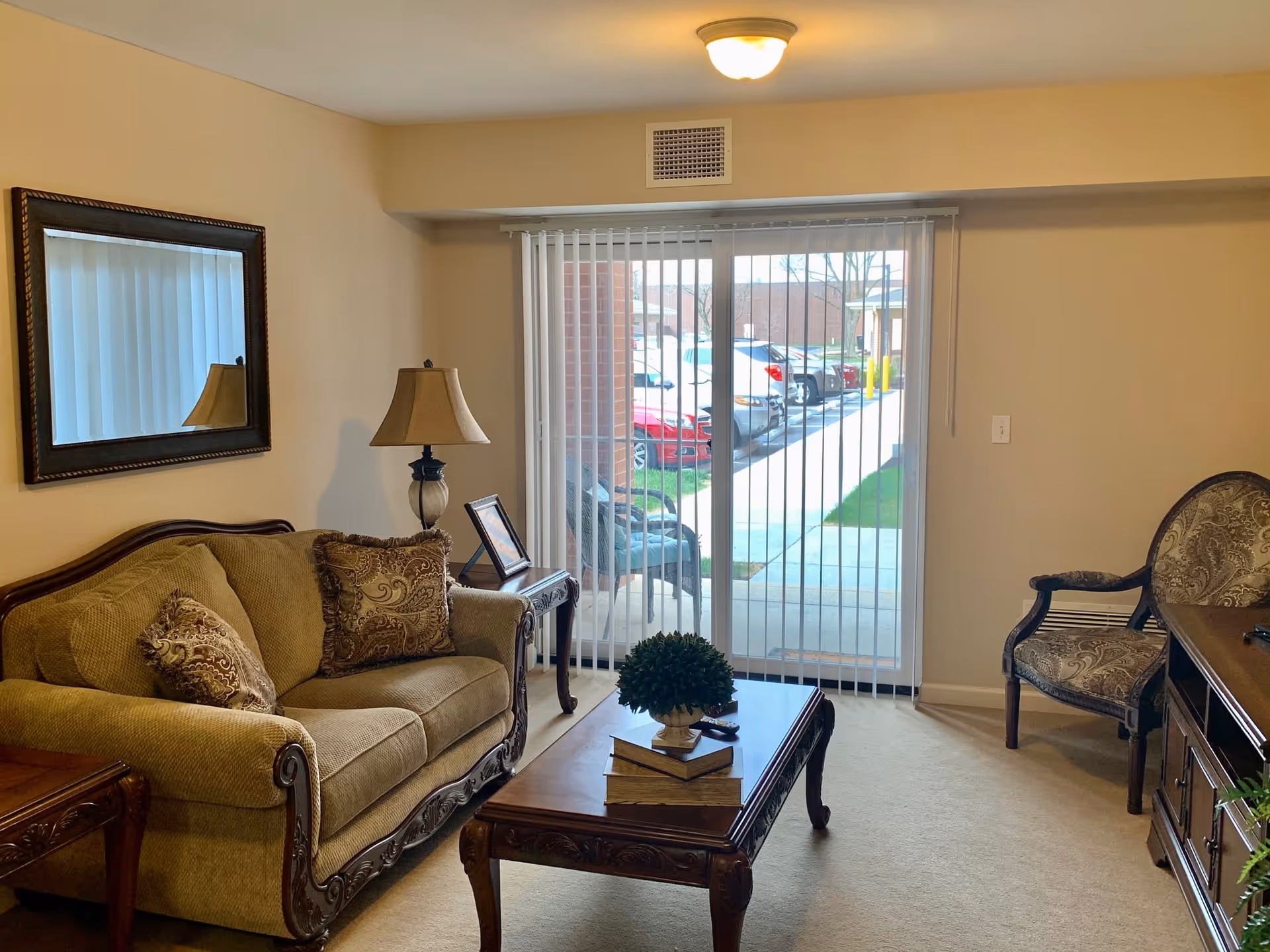 A cozy living room with a beige sofa adorned with patterned cushions, a wooden coffee table with books and a small plant, a side table with a lamp and a framed photo, an upholstered armchair, and a large mirror on the wall. Sliding glass doors with vertical blinds open to a view of a parking lot outside.