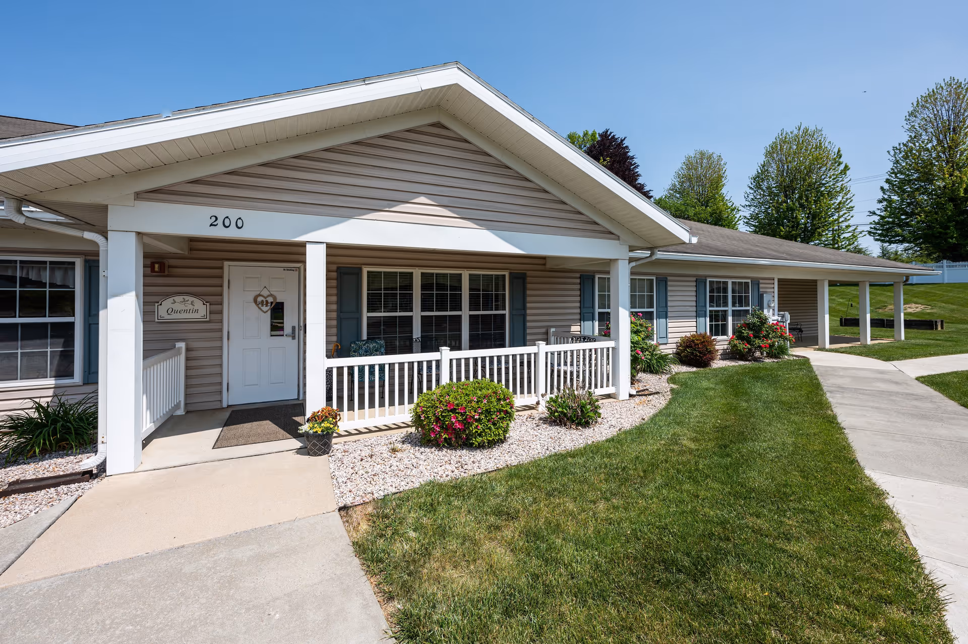 Exterior view of a single-story senior living community building with beige siding, white trim, and a covered porch area. The entrance door is labeled with the number 200 and a sign that reads 'Quentin'. There are bushes and flowers planted along the walkway and a well-maintained lawn under a clear blue sky.