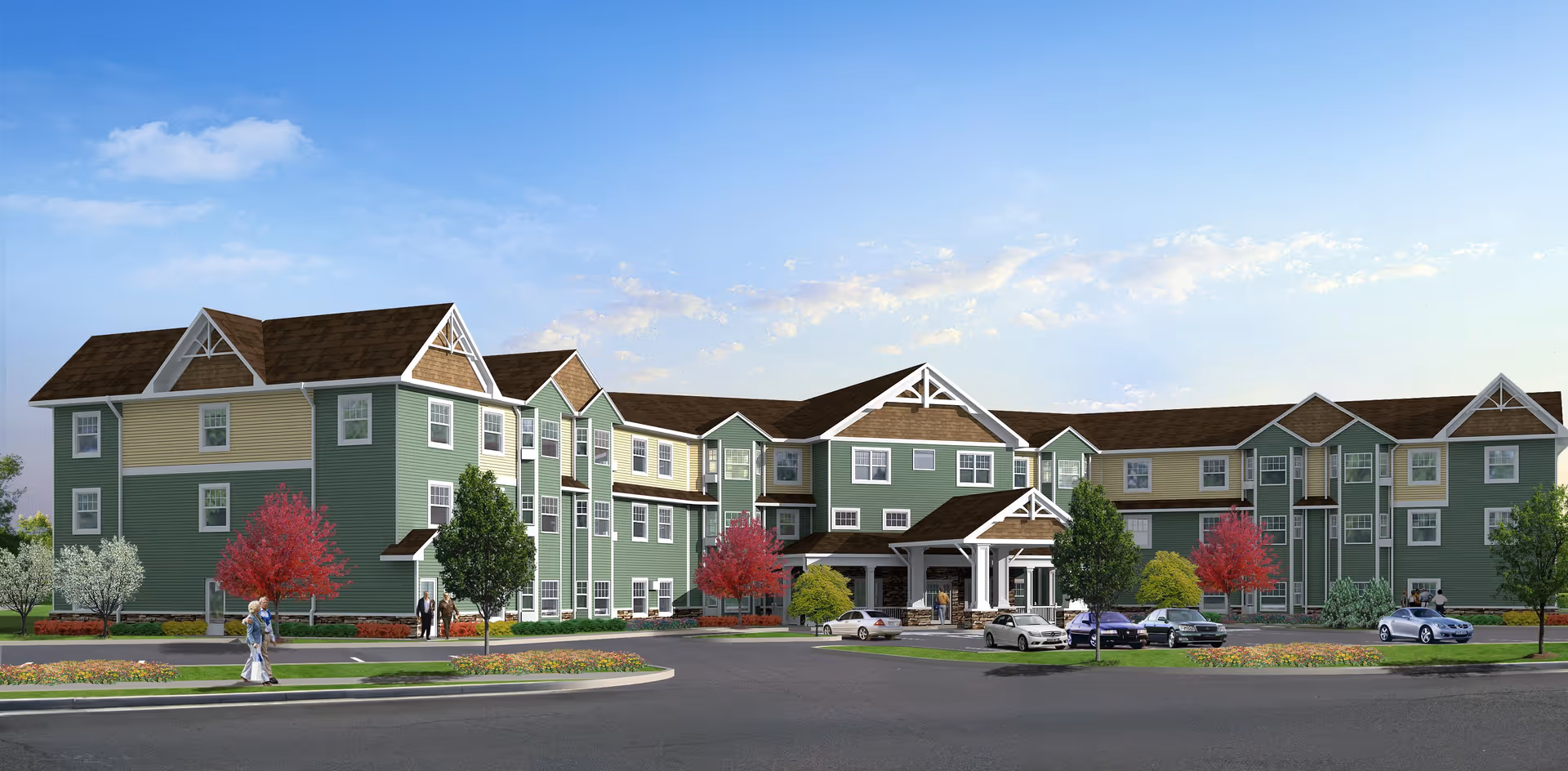 Exterior view of a large, three-story senior living facility building with green and beige siding, white trim, and brown roofs. The building has multiple windows and a covered entrance with a few cars parked in front. There are trees with red and green foliage and a clear blue sky with some clouds.