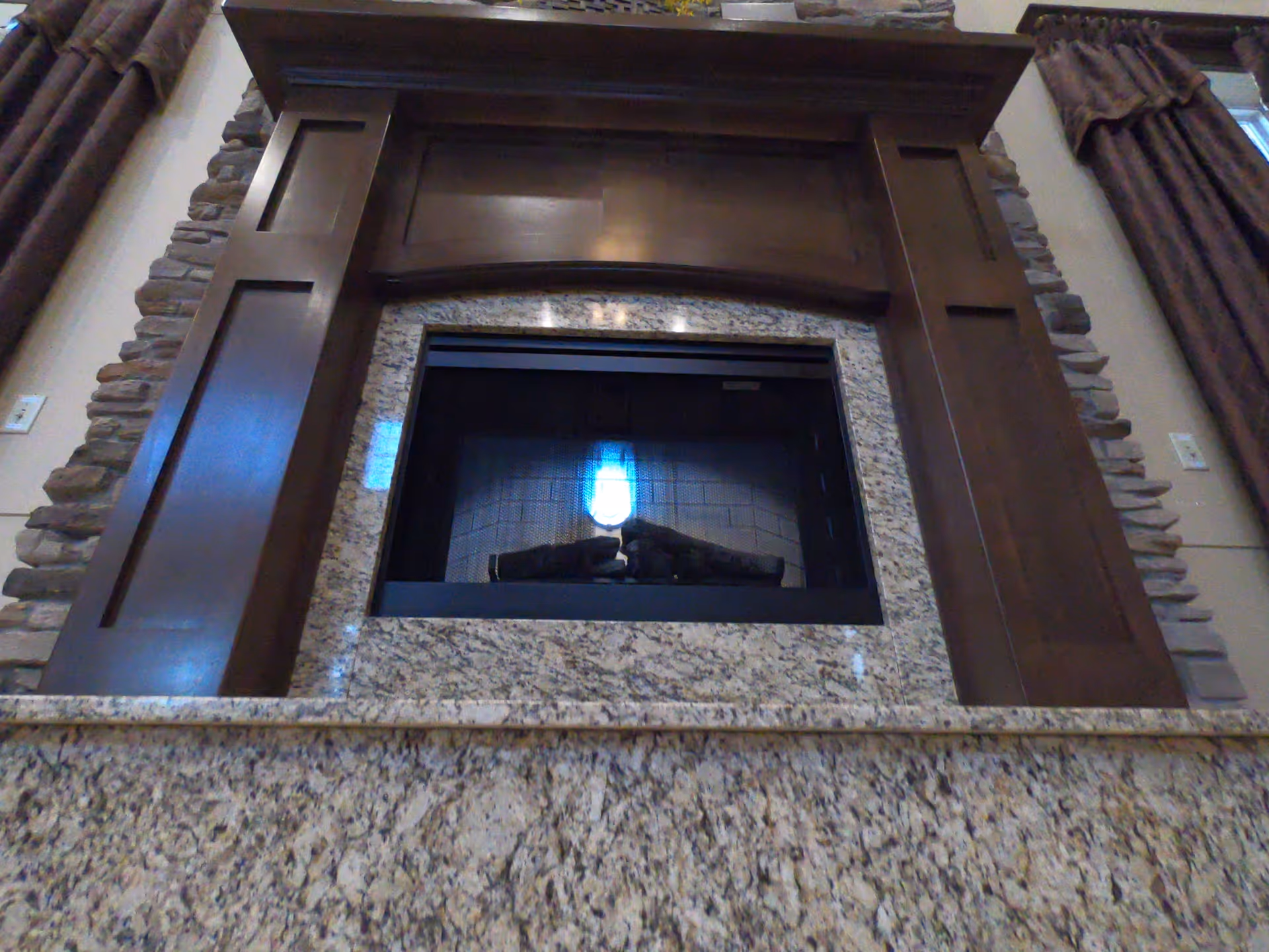 Close-up view of a fireplace with a granite surround and a wooden mantel. The fireplace has a glass front with logs inside and is flanked by stone accents on the wall. Brown curtains are partially visible on either side.