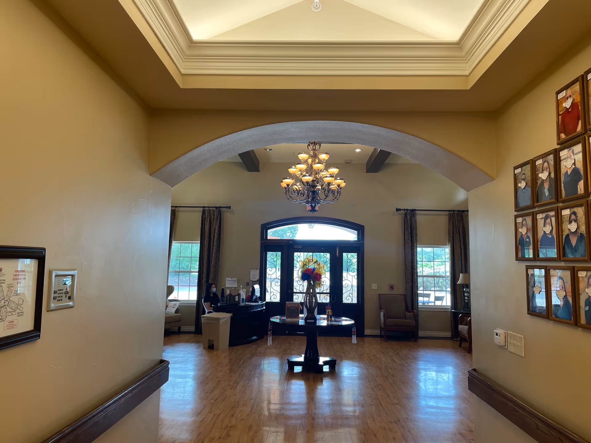 Interior view of a senior living facility lobby with a wooden floor, a round table with a flower vase in the center, a chandelier hanging from the ceiling, and a reception desk with a person wearing a mask seated behind it. There are framed portraits on the right wall and large windows with curtains on either side of the entrance door.