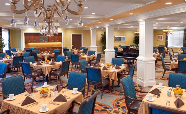 Bright, upscale dining room with many set tables, teal upholstered chairs, chandeliers, and a grand piano.