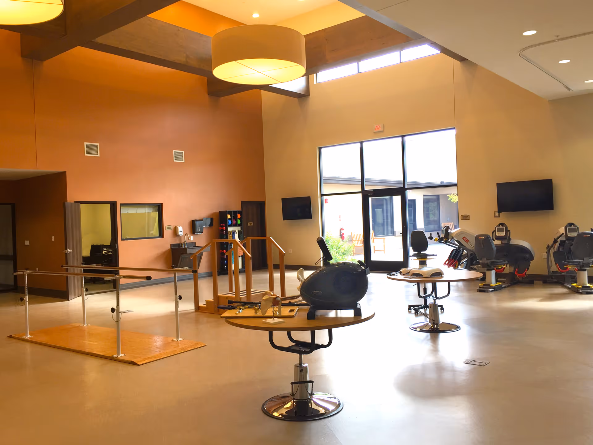 A spacious rehabilitation room with exercise equipment including stationary bikes, parallel bars for walking practice, and tables with therapy tools. The room has high ceilings, large windows letting in natural light, and warm-colored walls.