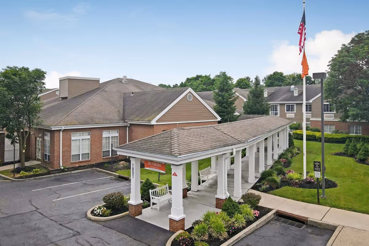 Covered porte-cochère with white columns and benches at the entrance of a brick senior living building, with lawns, flagpoles, and a parking area.