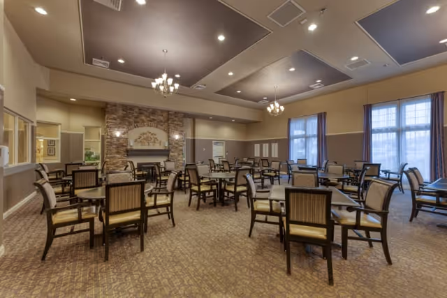 A spacious dining room with multiple square tables and cushioned chairs arranged neatly. The room features a high ceiling with recessed lighting and chandeliers, large windows with curtains allowing natural light, and a decorative stone fireplace with a floral arrangement above it. The carpet has a patterned design and the walls are painted in warm neutral tones.