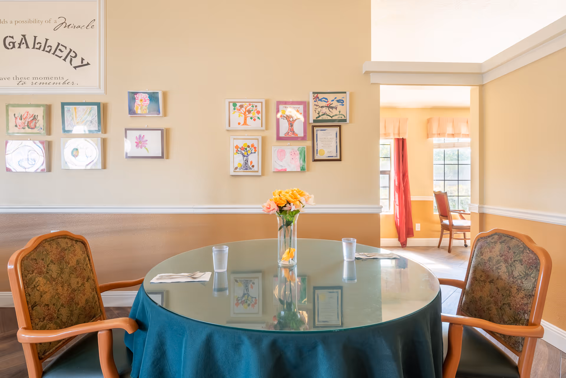 A round glass-top table covered with a teal tablecloth, set with two glasses of water and napkins, surrounded by two upholstered chairs with wooden arms. On the wall behind the table are several framed drawings and a sign that reads 'Gallery'. The room has warm beige walls with white trim, and an adjacent room with large windows and red curtains is visible.