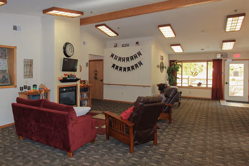 A cozy living room area in an assisted living facility featuring a maroon couch, two brown leather recliners, a small wooden bookshelf, a wall-mounted TV above an electric fireplace, and a large window with red curtains letting in natural light. The room has carpeted floors and ceiling lights with wooden beams.