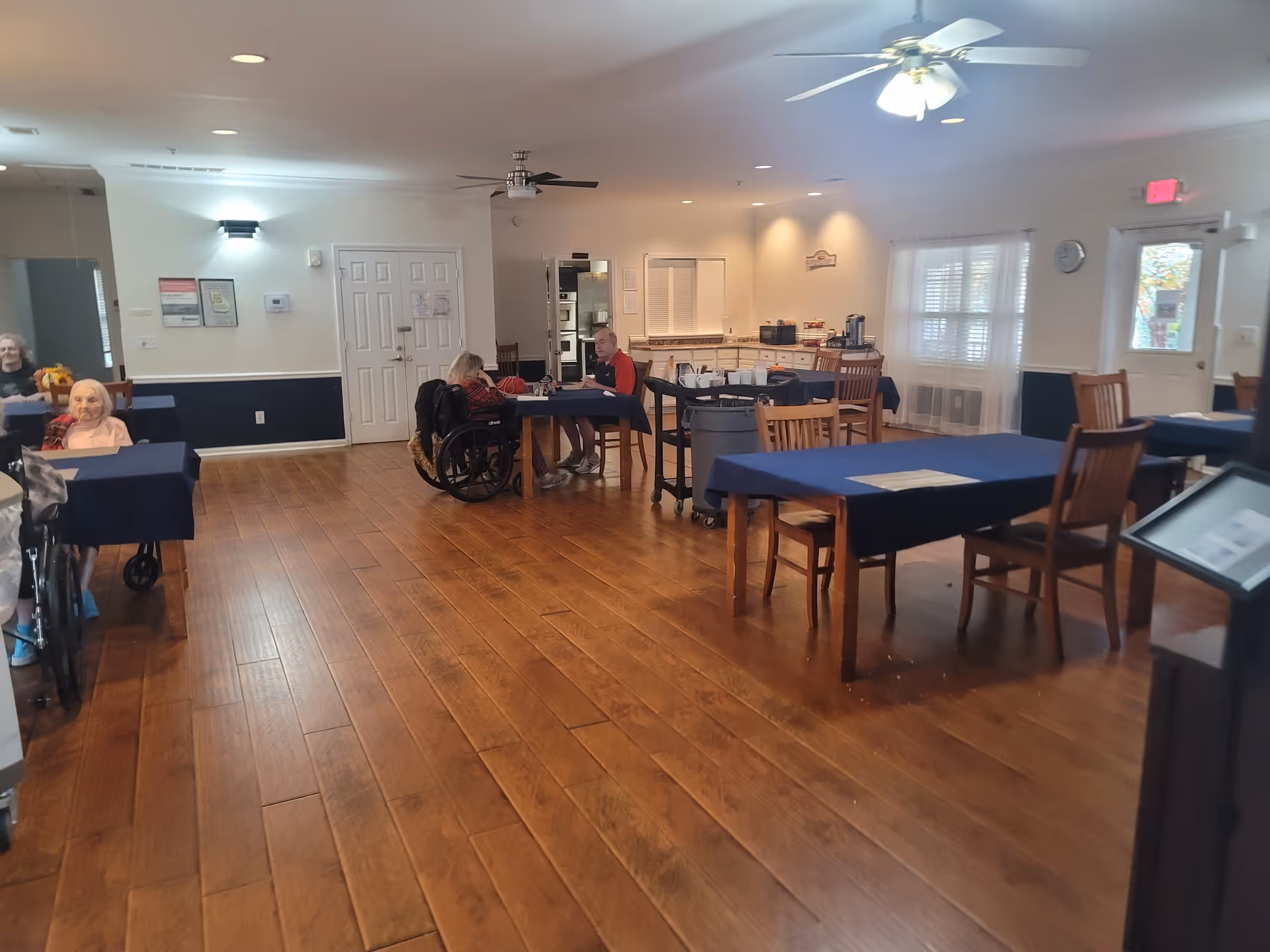 A spacious dining room in a senior living facility with wooden floors and several tables covered with blue tablecloths. There are a few elderly residents seated at the tables, some in wheelchairs. The room has ceiling fans, windows with white curtains, and a door leading outside. The back wall has a counter area with coffee and other supplies.