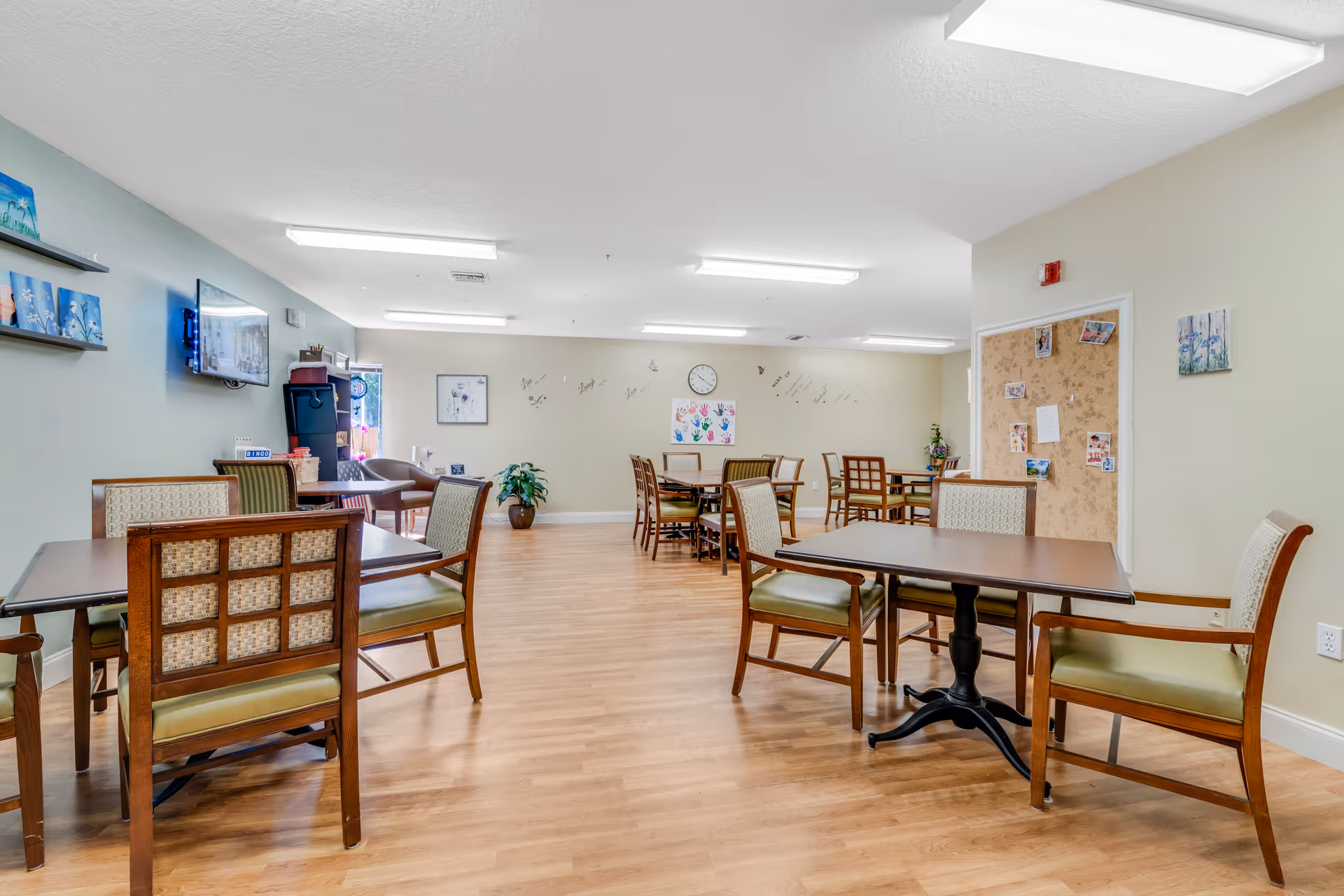 A bright and clean dining room with several wooden tables and chairs arranged neatly on a light wood floor. The walls are light-colored with some artwork and a bulletin board. A flat-screen TV is mounted on one wall, and there are fluorescent ceiling lights providing ample lighting.