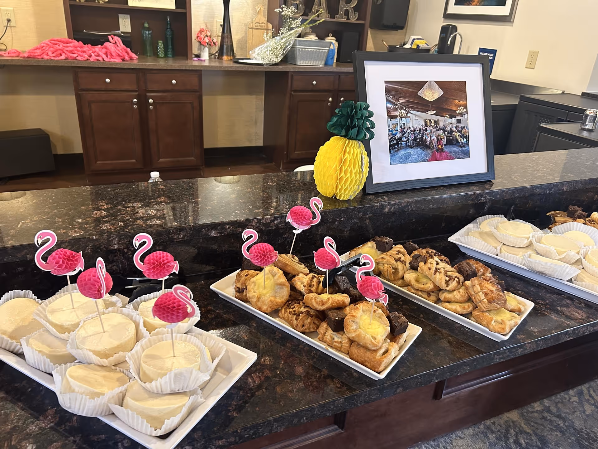 A countertop display of trays of pastries and desserts decorated with pink flamingo picks and a framed photo in a senior living facility.