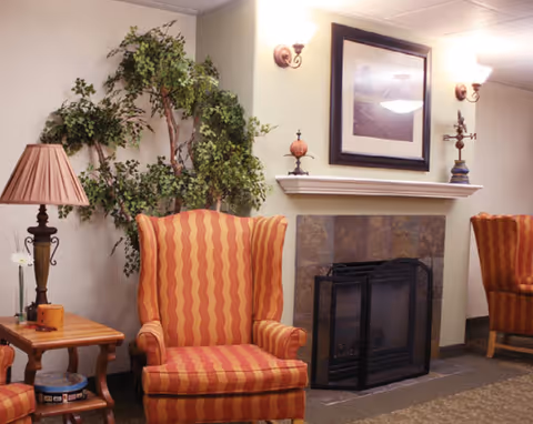 Cozy seating area with striped wingback chairs, a fireplace with framed art above, side tables, lamps, and a potted plant.