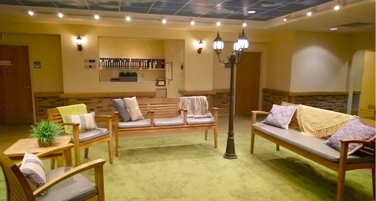 A cozy indoor seating area with wooden benches and chairs arranged around a vintage-style street lamp. The benches have cushions and throw blankets, and there is a small wooden table with a potted plant. The room has soft lighting, a green carpet, and a reception window with shelves and files in the background.
