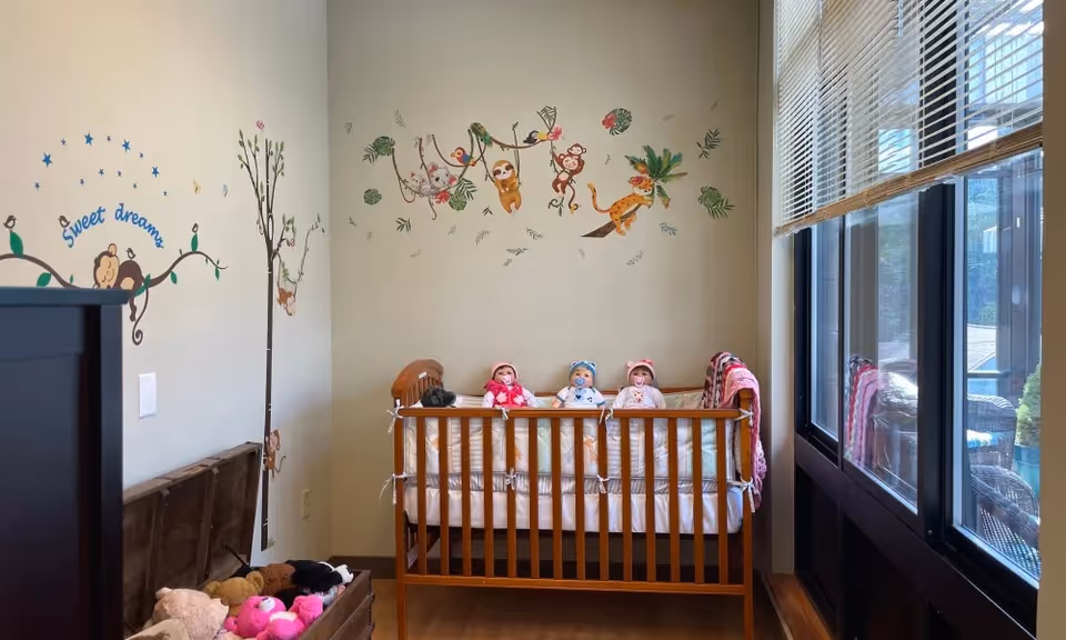 Small nursery with a wooden crib holding three dolls, jungle-themed wall decals, a trunk of stuffed animals, and large windows with blinds.