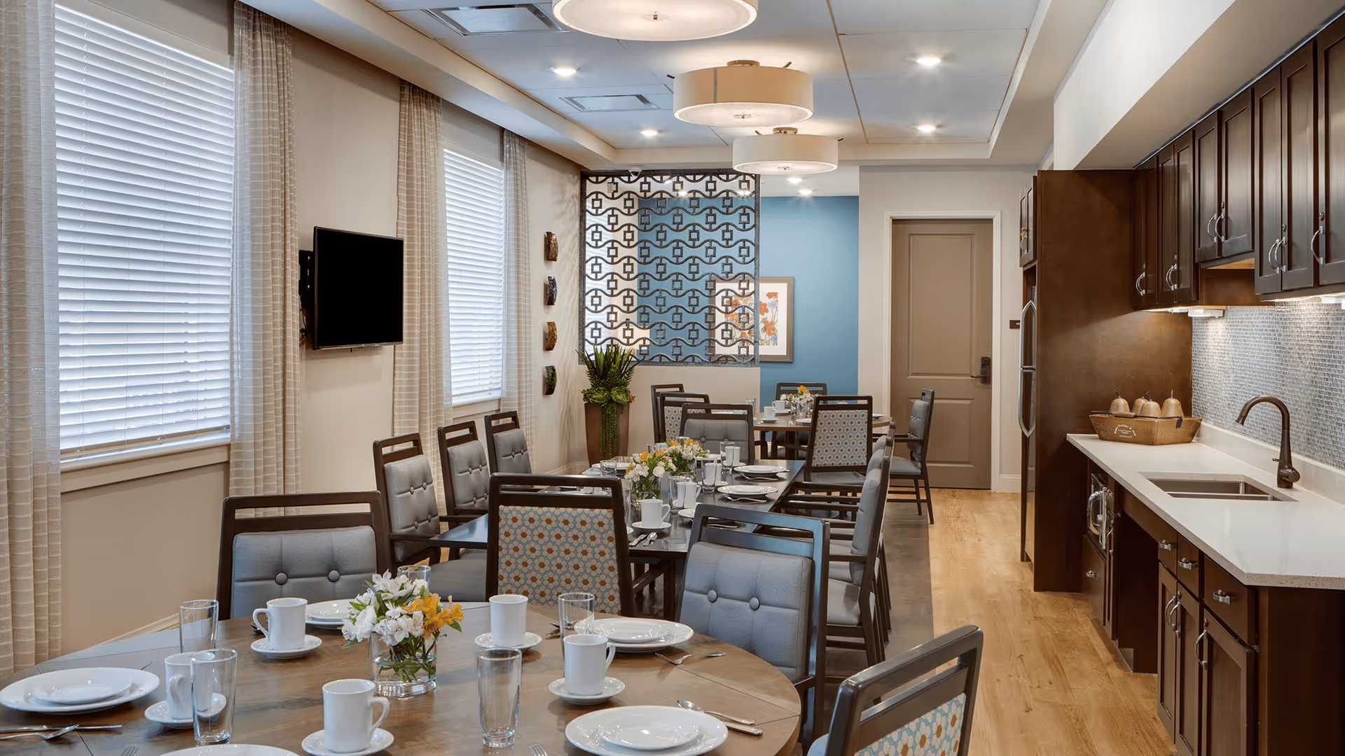 A dining room with multiple tables set for a meal, featuring plates, cups, glasses, and floral centerpieces. The room has large windows with blinds and curtains, a wall-mounted TV, and a kitchen area with dark wood cabinets, a sink, and a countertop. The walls are decorated with artwork and a patterned room divider.