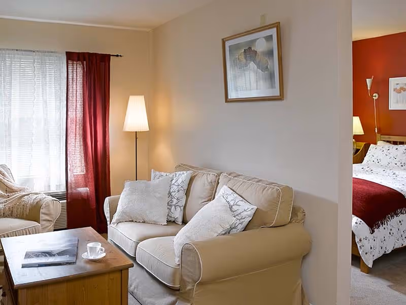 A cozy living room area with a beige loveseat adorned with white and patterned pillows, a wooden coffee table with a magazine and a cup on it, a floor lamp, and red curtains covering a window. Adjacent to the living room is a bedroom with a bed covered in white bedding with a red throw blanket and a bedside lamp.