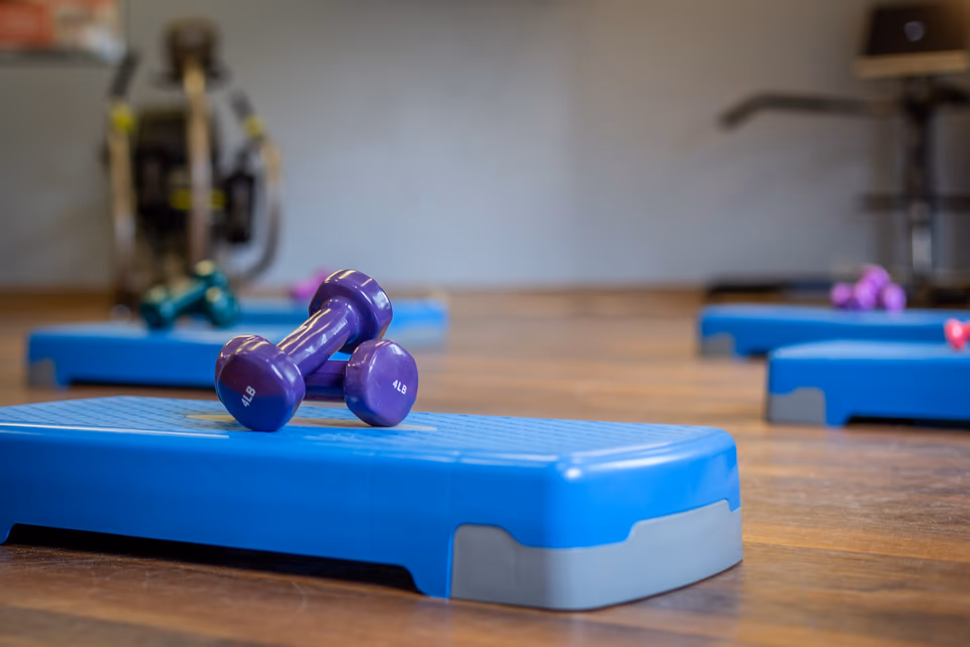 Close-up of a blue aerobic step platform with a pair of purple 4-pound dumbbells resting on top, set on a wooden floor in a fitness room with additional exercise equipment blurred in the background.