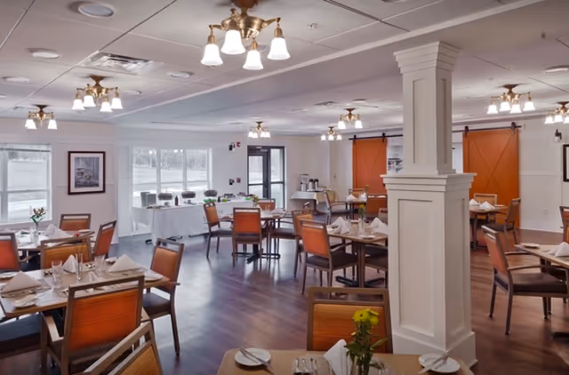 A spacious dining room with multiple tables and chairs arranged neatly. Each table is set with white napkins, plates, glasses, and cutlery. The room has wooden flooring, white walls, and several ceiling light fixtures. Large windows allow natural light to enter, and there are two orange sliding barn doors on one side. A buffet table with covered dishes is visible near the windows.