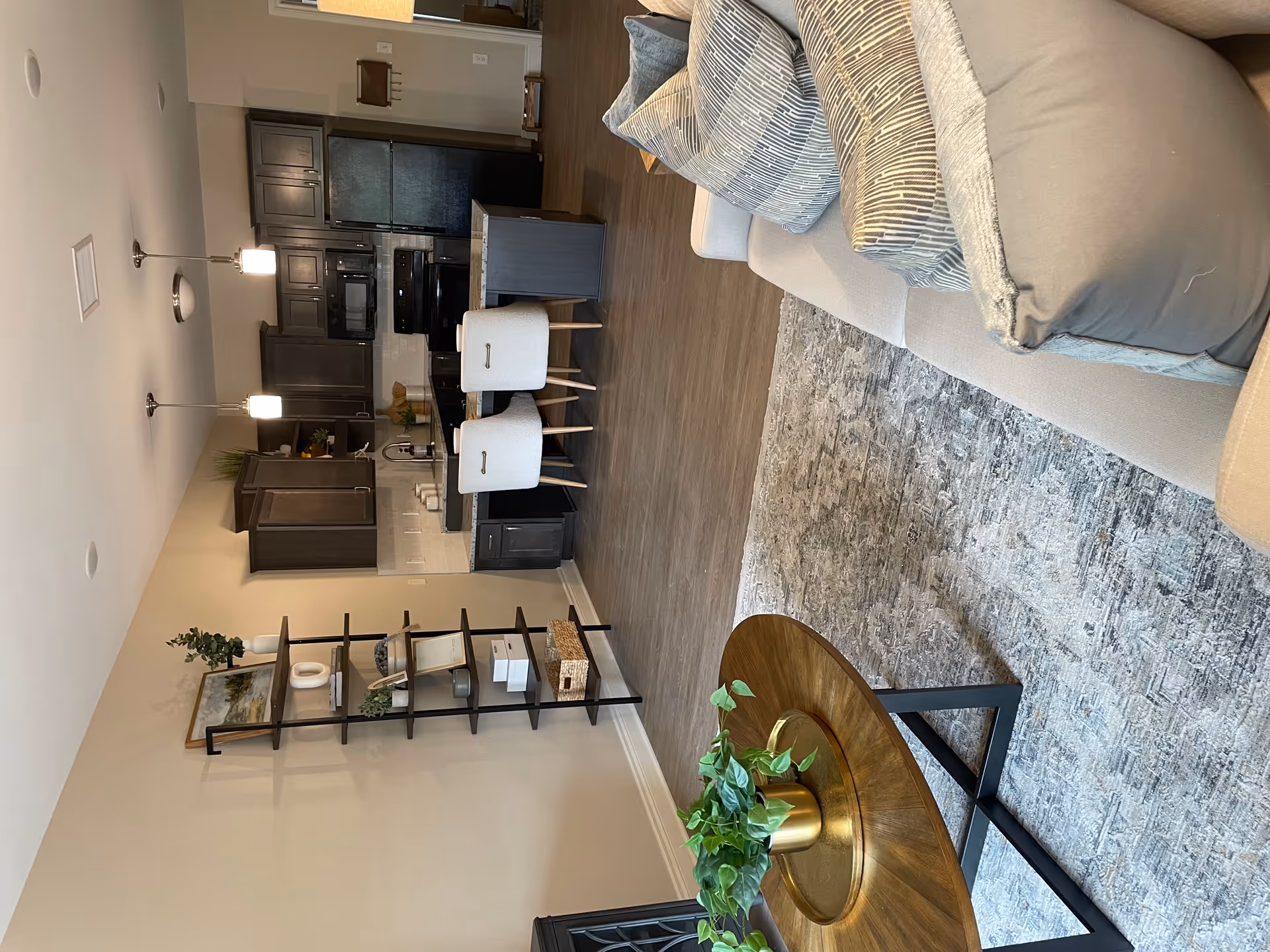 A modern senior living space featuring a cozy living room with a light gray sofa adorned with patterned pillows, a wooden coffee table with a green plant in a gold pot, and a textured area rug. In the background, there is an open kitchen with dark wood cabinets, black appliances, a granite countertop island with two white upholstered chairs, and pendant lighting overhead. A black metal shelving unit with decorative items is positioned against the beige wall.