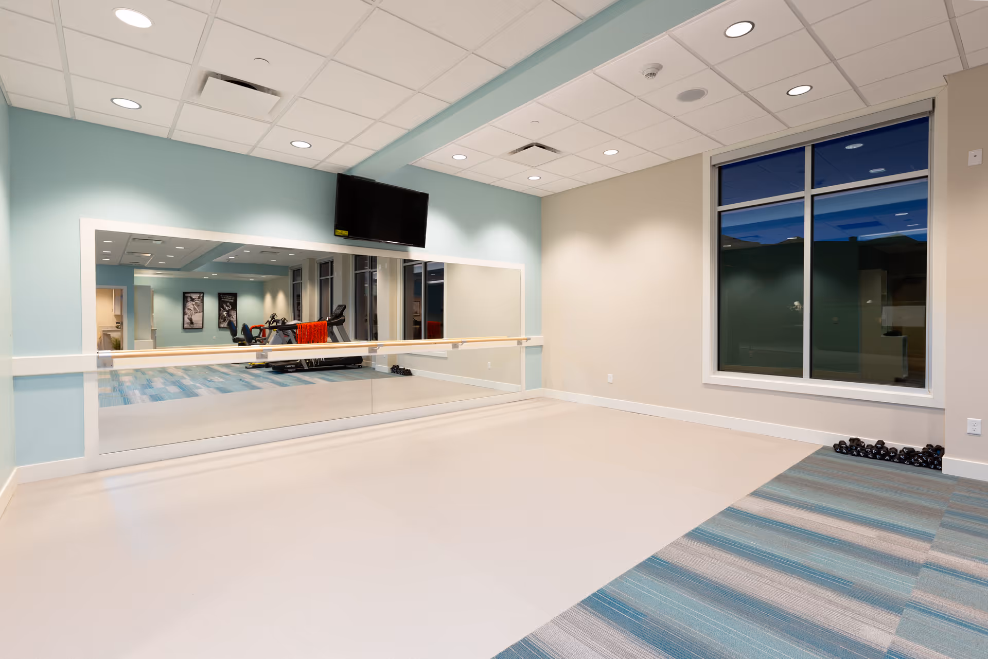 A spacious exercise room with a large wall mirror, a mounted flat-screen TV, and a ballet barre. The room has light blue and beige walls, a window, and a section of carpeted floor with blue and gray stripes. In the reflection of the mirror, exercise equipment including a treadmill and stationary bike are visible.