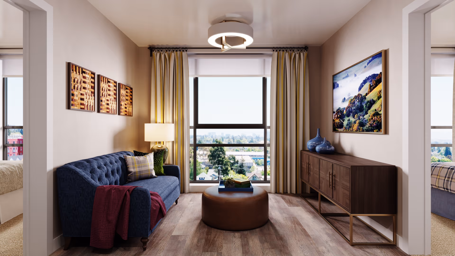 A cozy living room with a blue tufted sofa adorned with pillows and a red throw blanket, a round brown ottoman in the center, a wooden sideboard with blue vases, and a large window with curtains offering a view of trees and buildings outside. There are two doorways on either side leading to bedrooms.