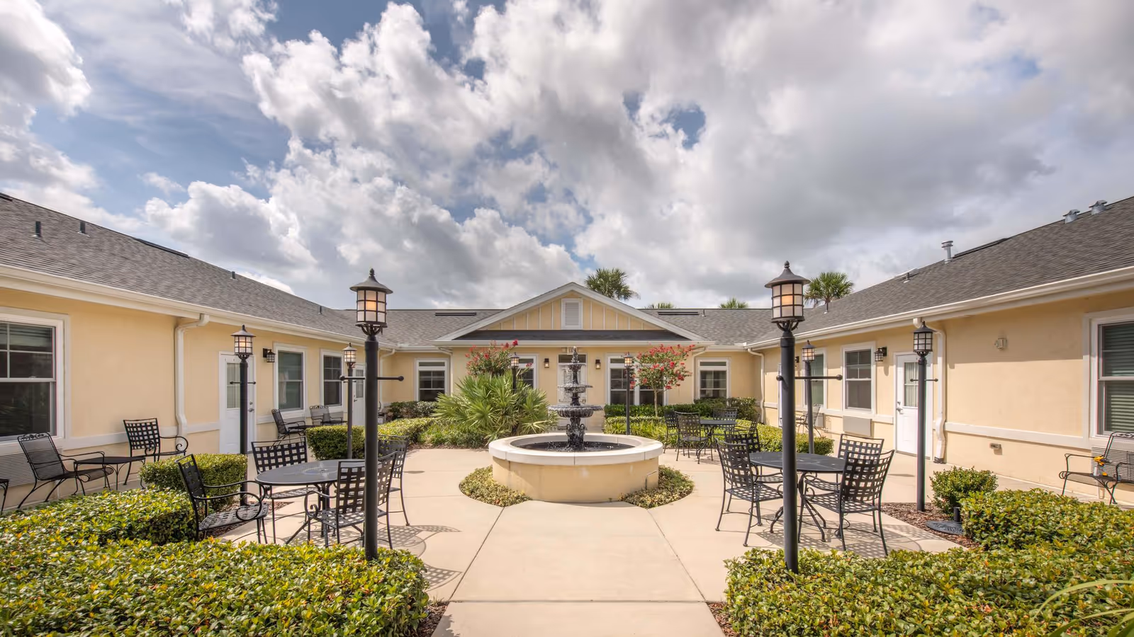 Outdoor courtyard area at Benton House of Clermont featuring a central water fountain surrounded by black metal tables and chairs, lamp posts, and beige buildings with windows and doors under a partly cloudy sky.