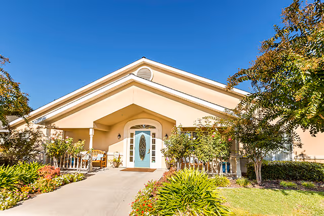 Front exterior view of a single-story building with a peaked roof, a light beige facade, and a teal front door with decorative glass. The entrance is flanked by white columns and surrounded by well-maintained landscaping including bushes, flowers, and trees under a clear blue sky.
