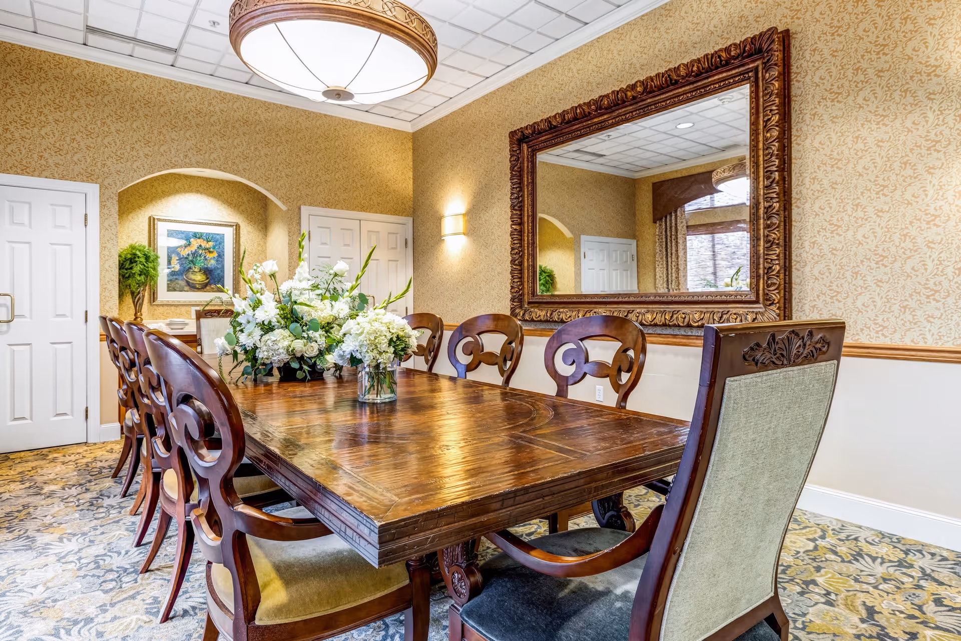 A formal dining room with a large wooden table surrounded by ornate wooden chairs with upholstered seats. The table is decorated with two floral arrangements featuring white flowers and greenery. The room has patterned wallpaper, a large decorative mirror on one wall, a framed painting, and a potted plant in a recessed alcove. The ceiling has a large round light fixture, and the floor is covered with a patterned carpet.