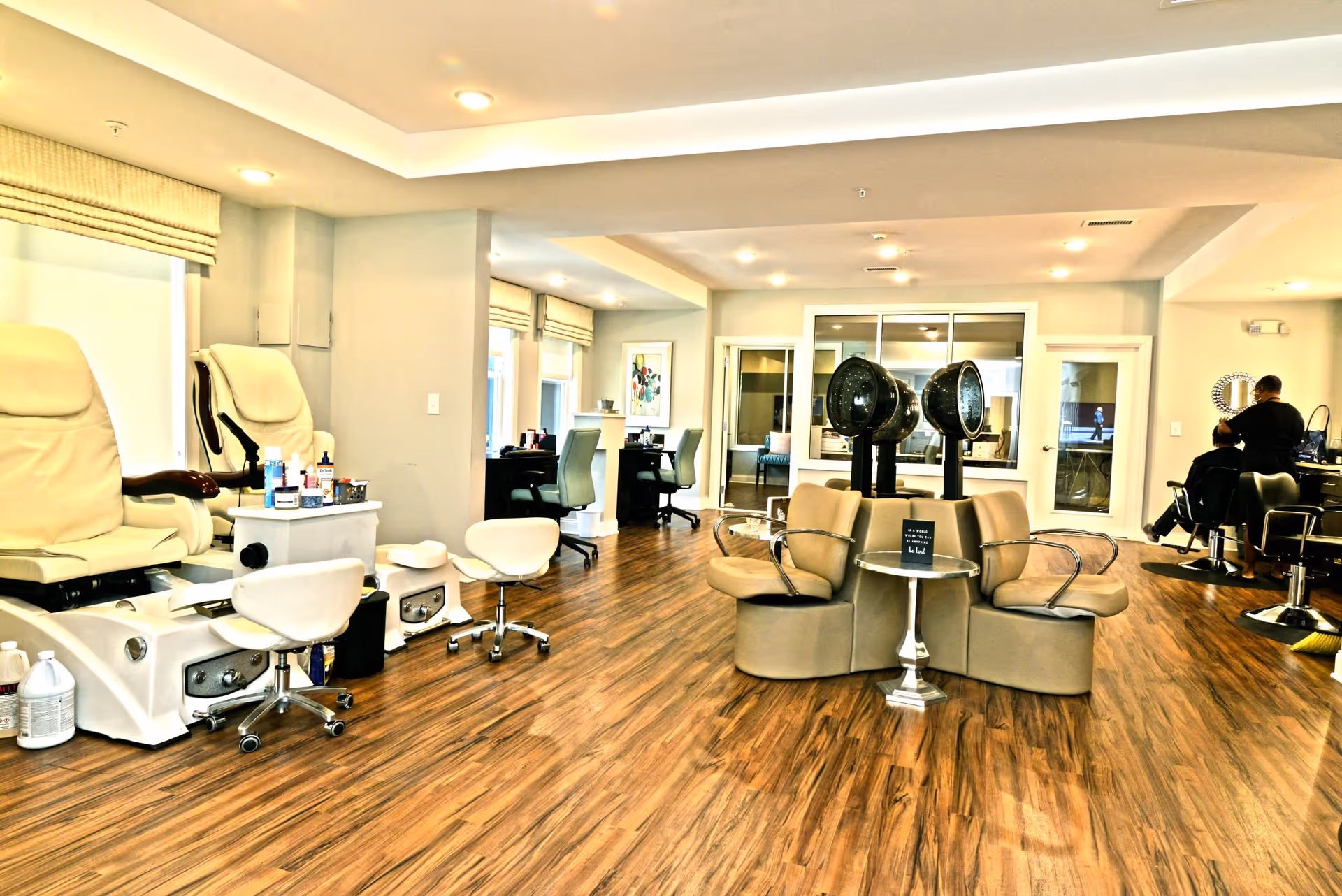 Interior view of a salon area in a senior living facility with pedicure chairs on the left, hair drying chairs in the center, and hair styling stations on the right. The room has wooden flooring, beige walls, and recessed ceiling lights.