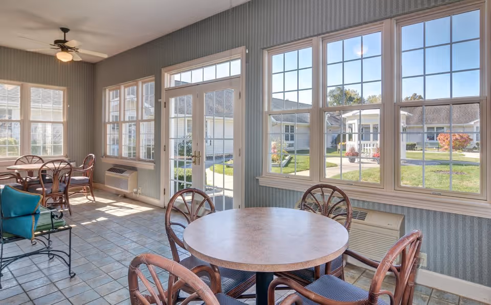 A bright sunroom or common area with multiple round tables and wicker chairs. Large windows and a glass door provide a clear view of a courtyard with grass, pathways, and other buildings. The room has tiled flooring, a ceiling fan with a light, and striped wallpaper.