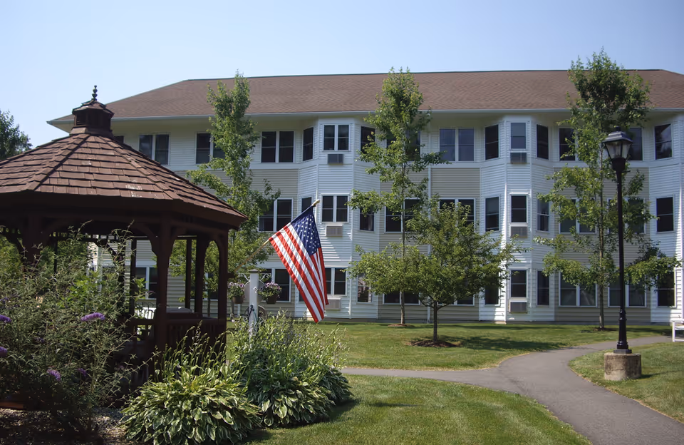 Front exterior of a multi-story white retirement community building with a gazebo, American flag, lawn, and walkway.