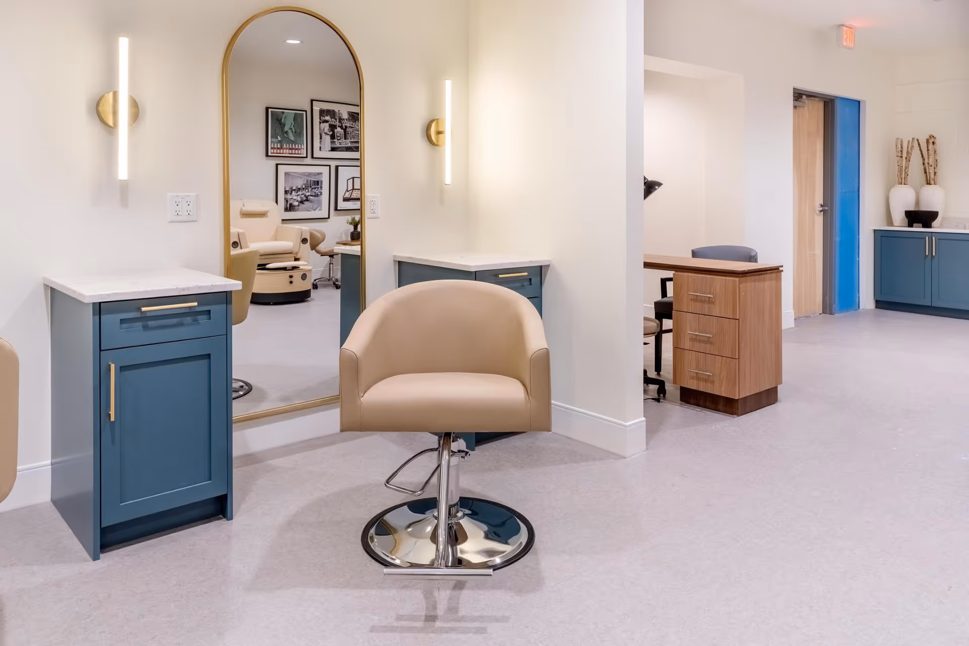 A bright salon-style interior with a beige styling chair in front of an arched mirror, blue cabinets, and wall sconces.