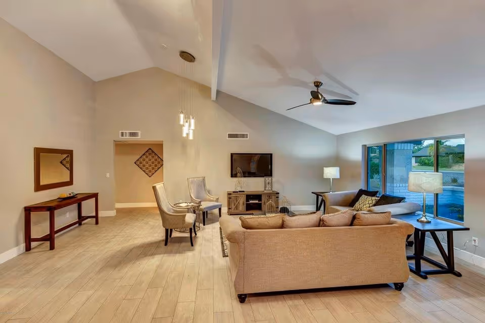 A spacious living room with beige walls and light wood flooring. The room features a beige sofa and two beige armchairs arranged around a wooden TV stand with a flat-screen TV mounted on the wall. There are two side tables with lamps, a ceiling fan with light, and a large window showing an outdoor view.