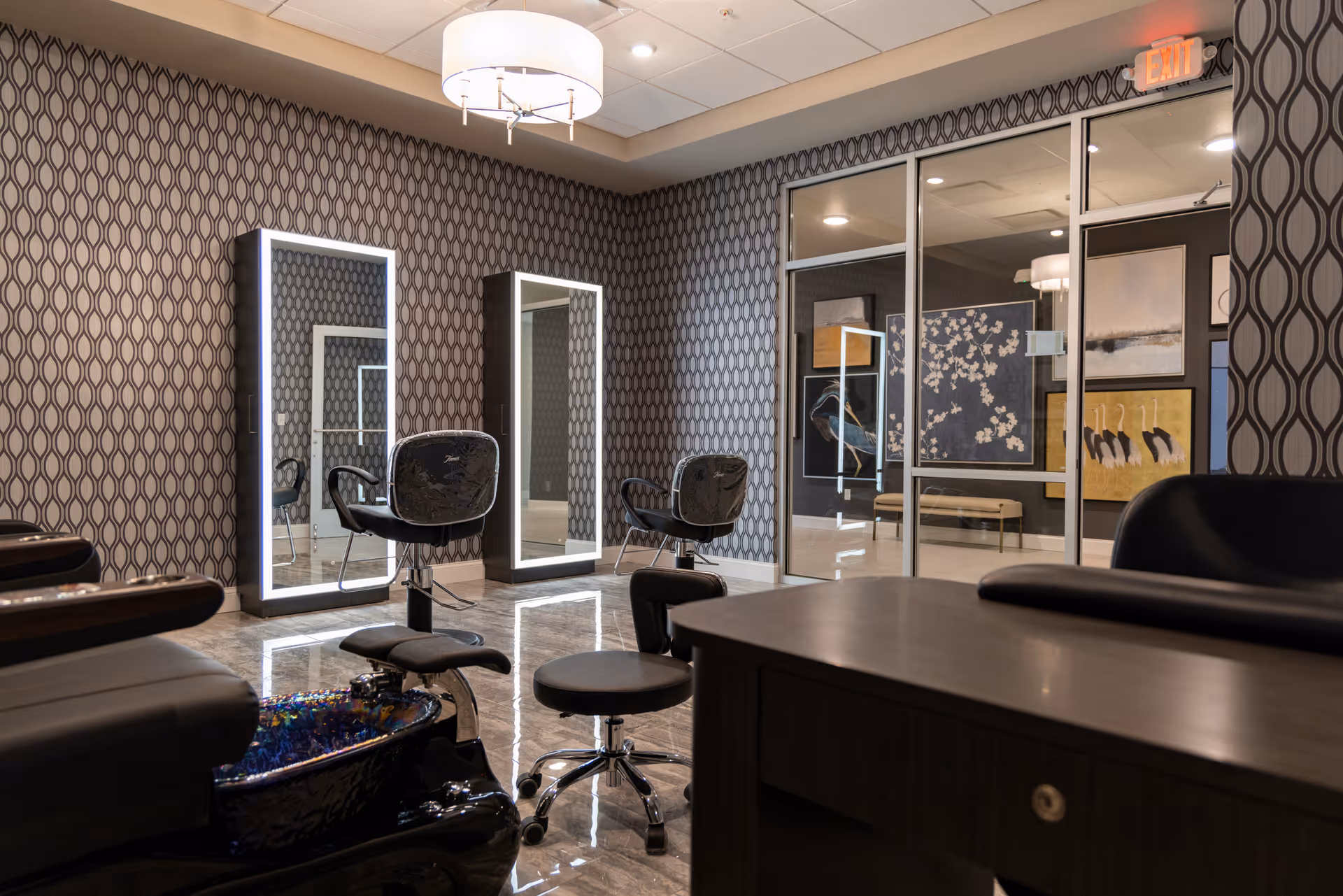 Interior view of a stylish salon area with two illuminated mirrors mounted on a patterned wall, two black salon chairs, a black pedicure chair with a footbath, a small rolling stool, and a dark wooden desk in the foreground. The room has a modern ceiling light fixture and glass doors leading to another room with framed artwork on the walls.