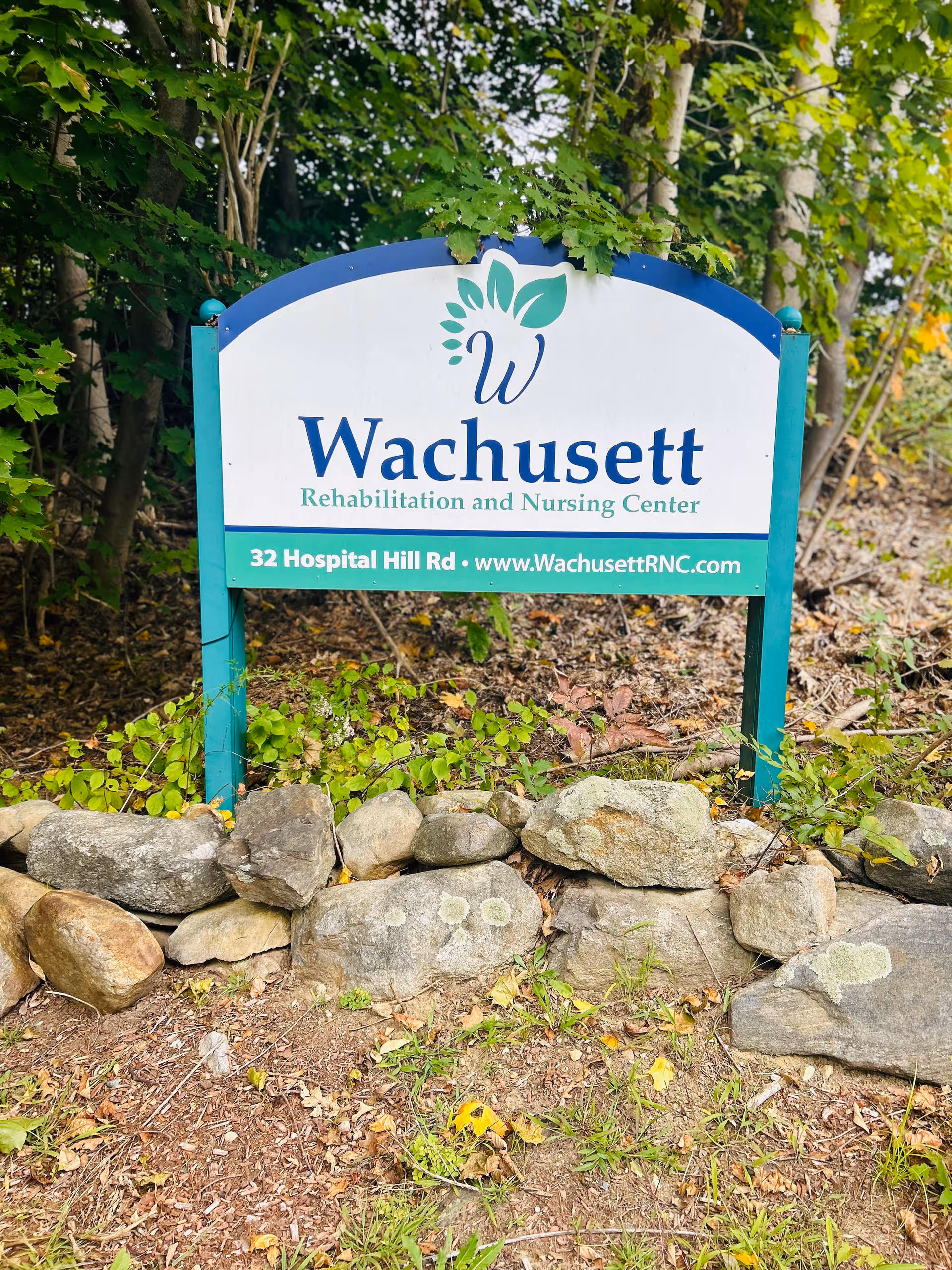 Outdoor sign reading "Wachusett Rehabilitation and Nursing Center" mounted among trees and rocks.