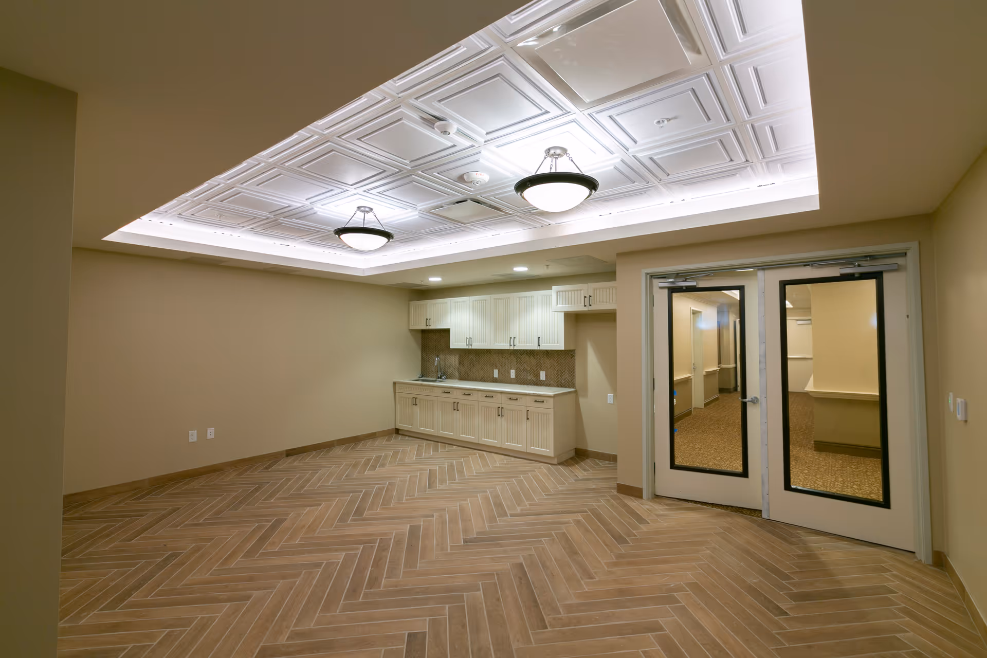 Empty common room with a built-in kitchenette, decorative recessed ceiling lights, and double glass doors.
