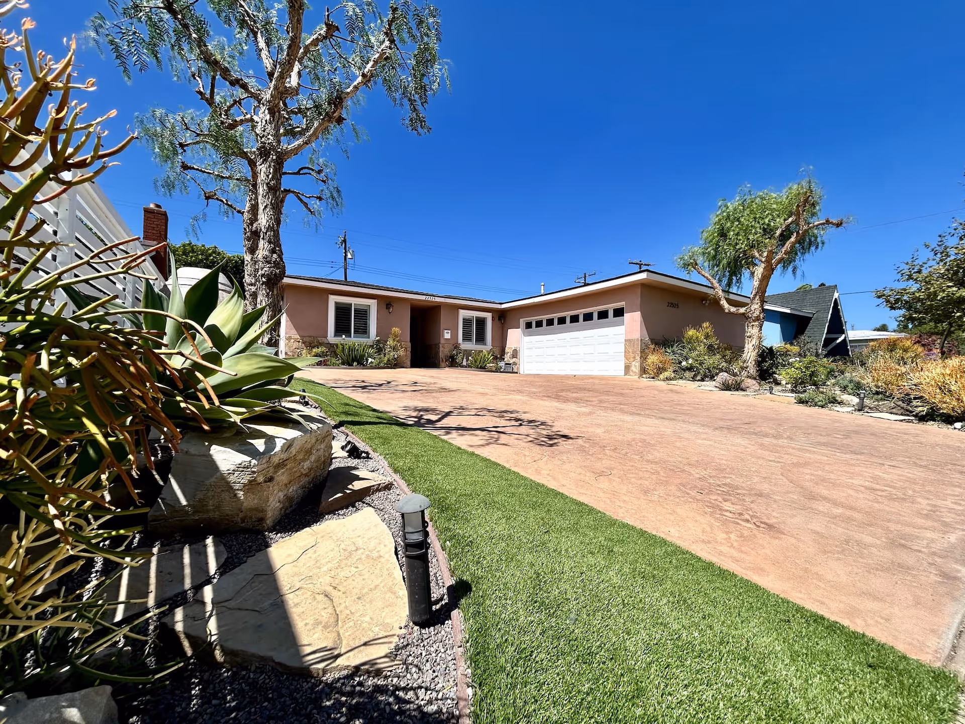 Exterior view of a single-story residential building with a wide driveway, a white garage door, and a front entrance. The yard features desert landscaping with various plants, rocks, and a tree under a clear blue sky.