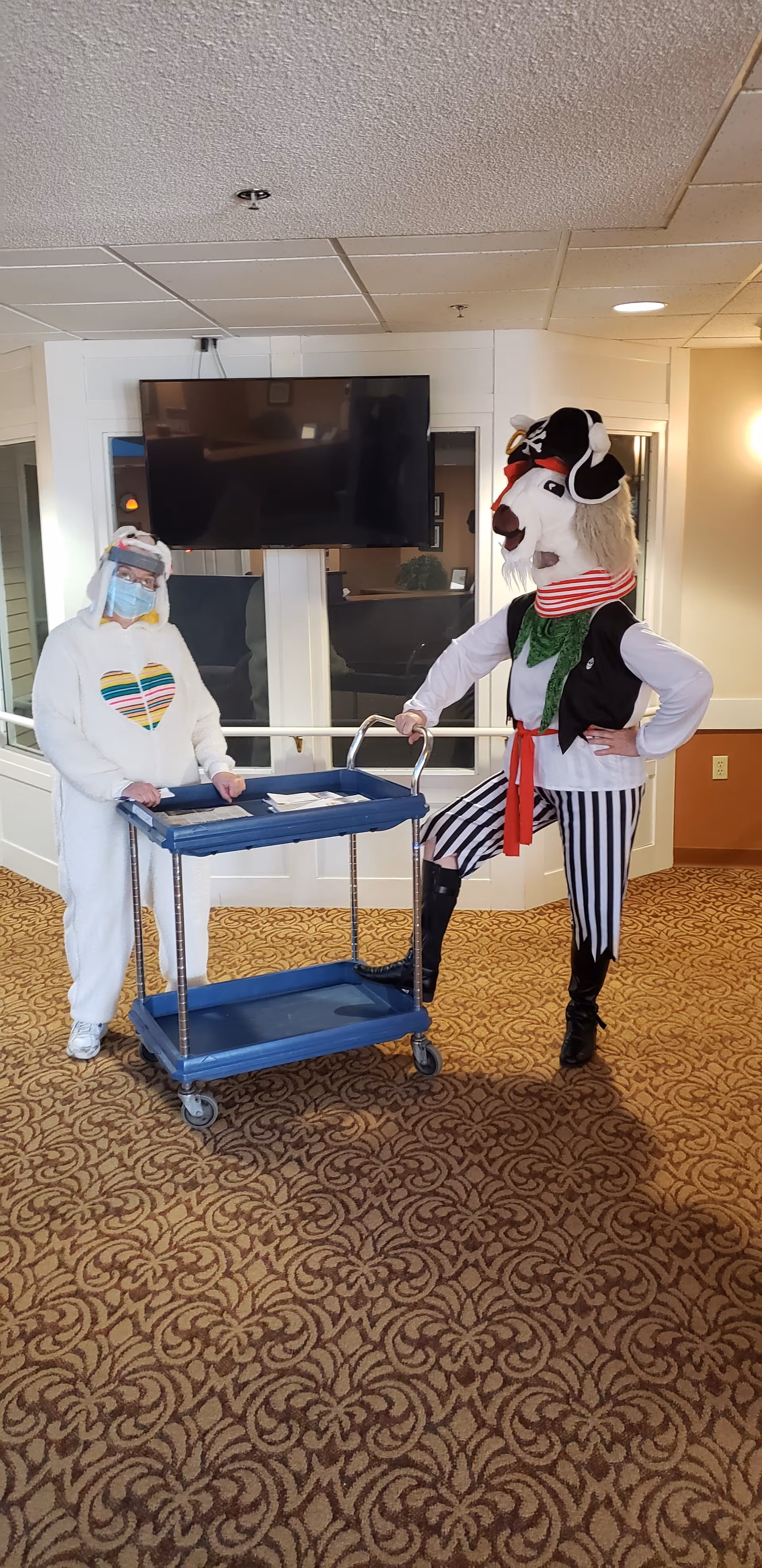 Two people in costumes stand indoors on a patterned carpet. One person is dressed in a white animal costume with a heart design on the chest and a face shield, standing behind a blue cart. The other person is dressed as a pirate with a large animal head, striped pants, a green scarf, and a black vest, standing with one foot on the cart handle. A flat-screen TV is mounted on the wall behind them.
