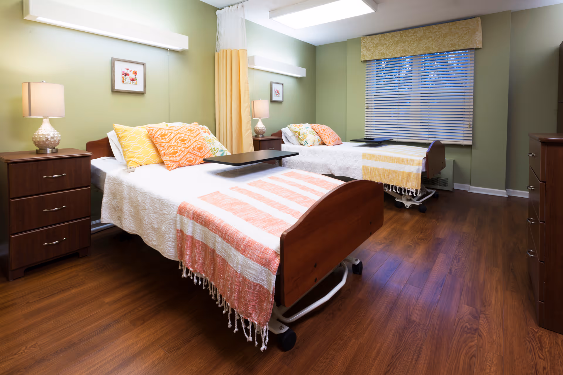 Shared bedroom with two hospital-style single beds, colorful pillows and throws, bedside tables with lamps, a window with blinds, and wood flooring.