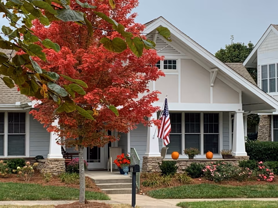 Front exterior view of a residential building with a porch decorated with pumpkins and an American flag. There is a tree with bright red autumn leaves in the front yard, along with green grass and various shrubs and flowers.