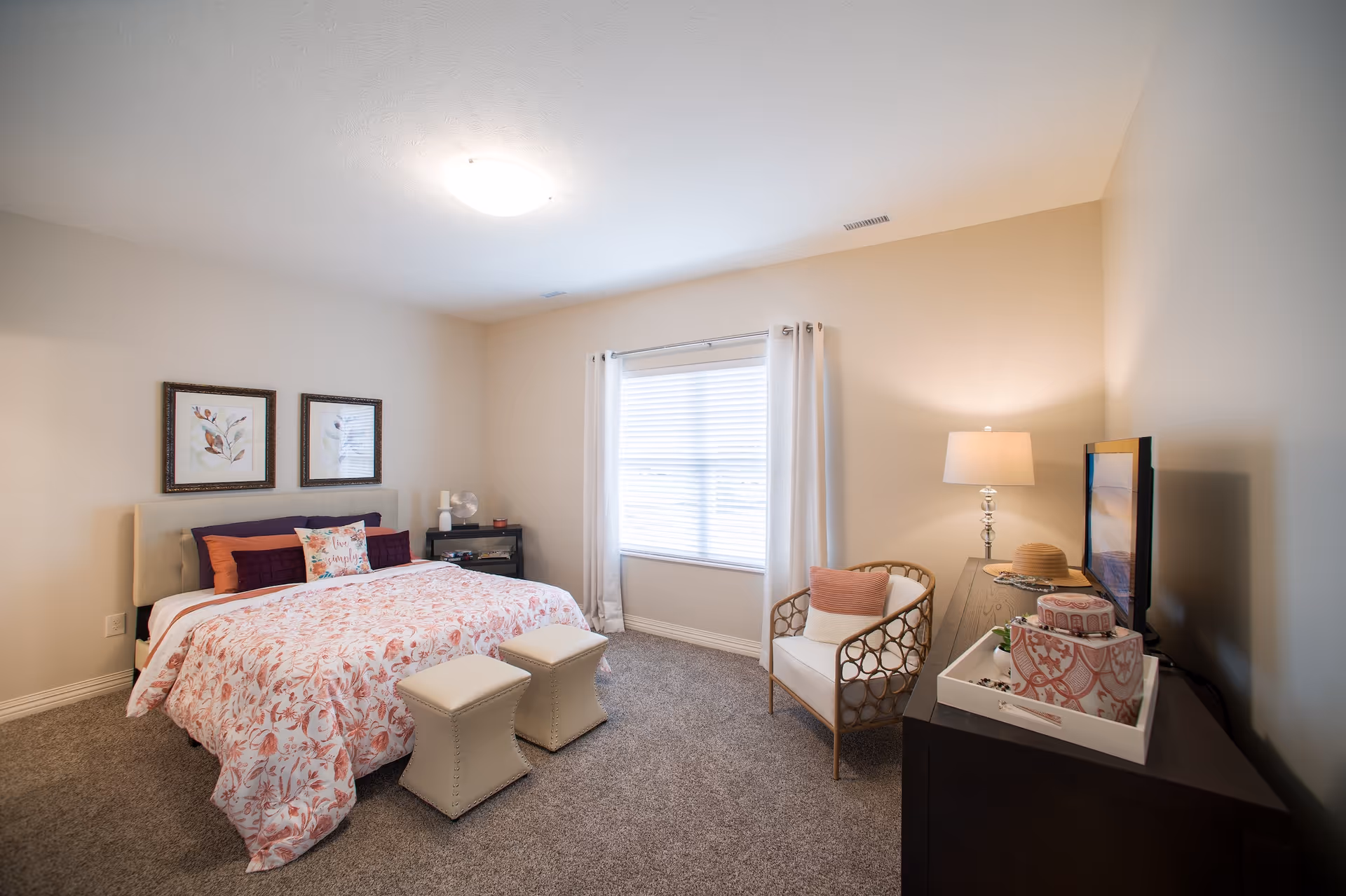 Bright furnished bedroom with a bed dressed in floral bedding, two small ottomans at the foot, an armchair by a window, and a dresser with a TV and lamp.