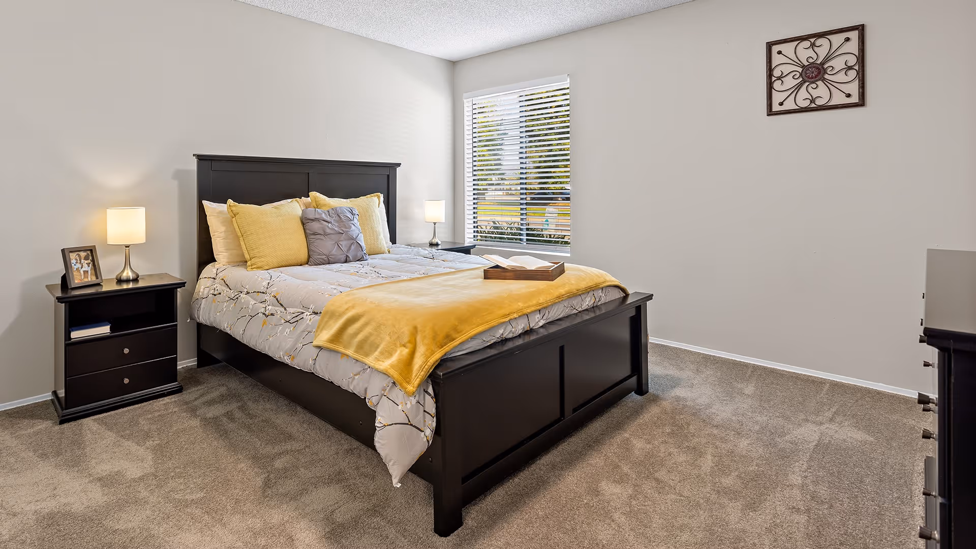 A neatly made bed with a dark wooden frame and headboard in a bedroom. The bed has a gray and white patterned comforter, yellow and gray pillows, and a yellow throw blanket. There are two bedside tables with lamps on each side of the bed, a framed photo on the left table, and a window with blinds letting in natural light. The room has beige carpet and light-colored walls with a decorative wall hanging.