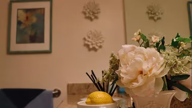Close-up view of a bathroom countertop with a decorative flower arrangement in a vase, a lemon placed on a small dish, and a toothbrush holder with toothbrushes. The background shows a towel hanging on a rack, a framed picture, and two decorative wall hangings.