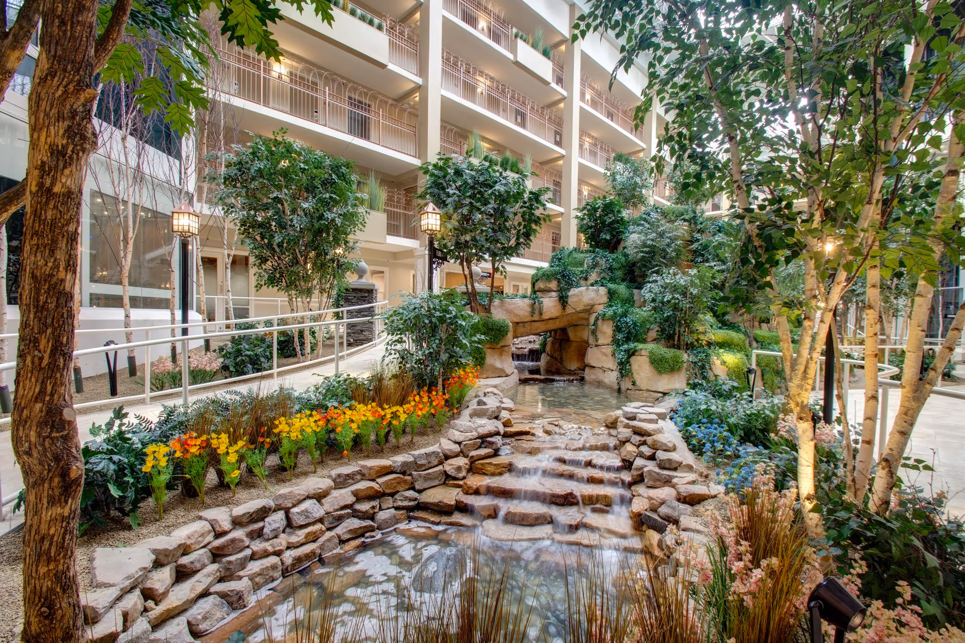 Indoor atrium featuring a terraced rock waterfall and pond surrounded by lush plants and interior balconies.