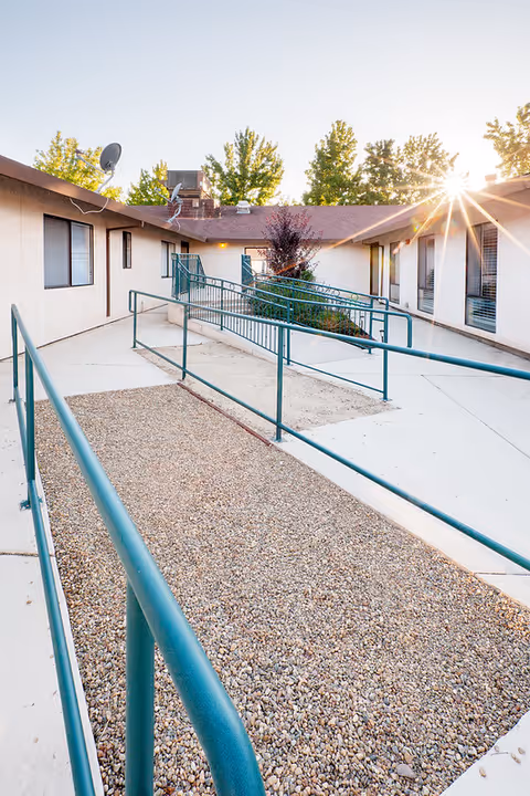 Outdoor courtyard area of a care facility with a gravel pathway and green metal handrails leading to building entrances. The sun is shining brightly over the roof, and there are trees in the background.
